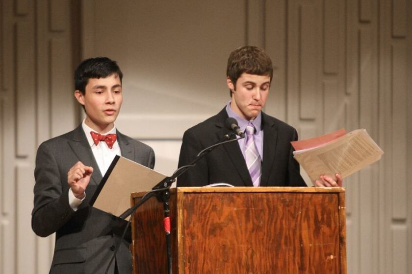 Two students at a wooden podium with a microphone, presenting in formal attire during a speech or debate exchange