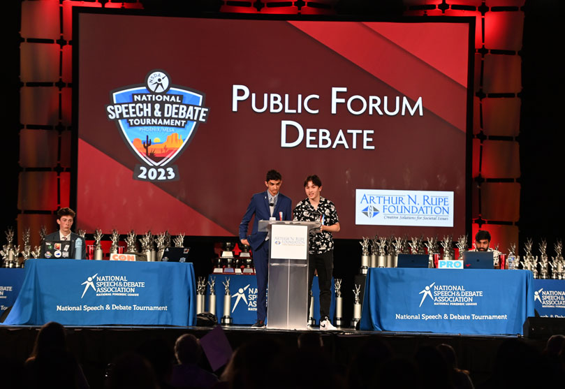 Public Forum debate on stage at the National Speech & Debate Tournament, with pro and con tables and event branding