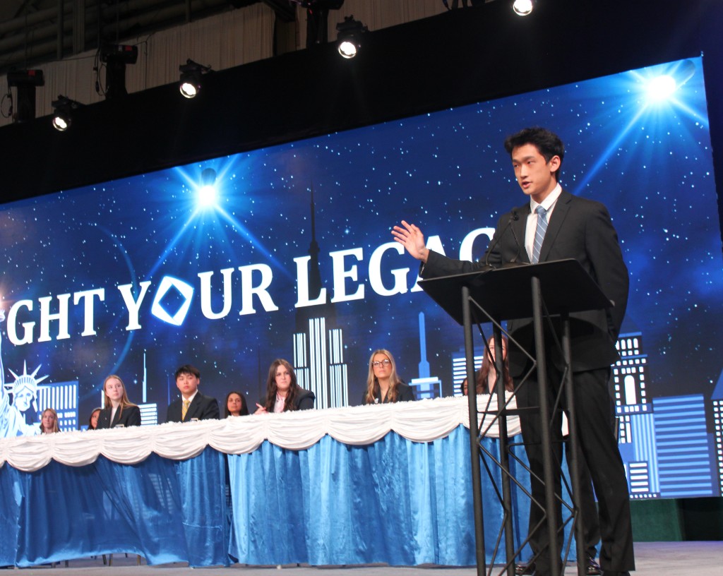 Student delivering a speech from a podium on a large stage with a professional panel and event backdrop