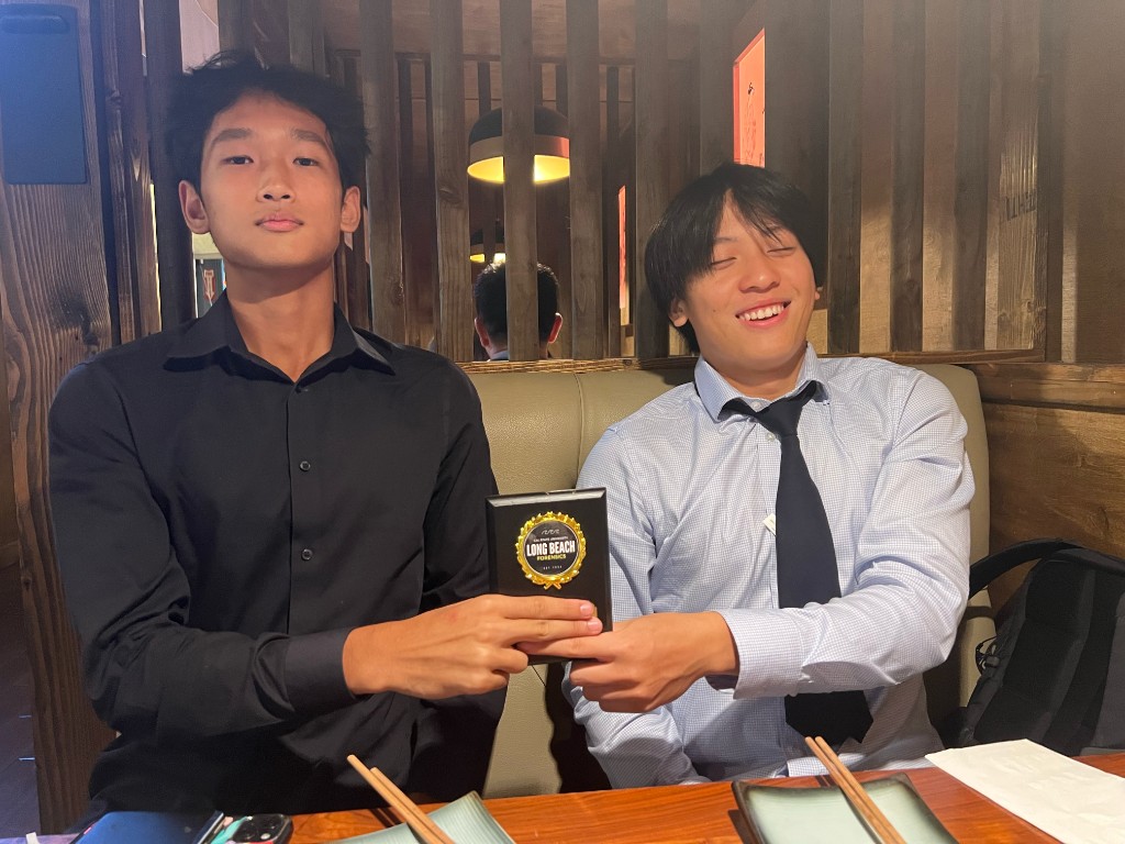 Two students at a restaurant table holding a Cal State Long Beach Forensics award plaque together