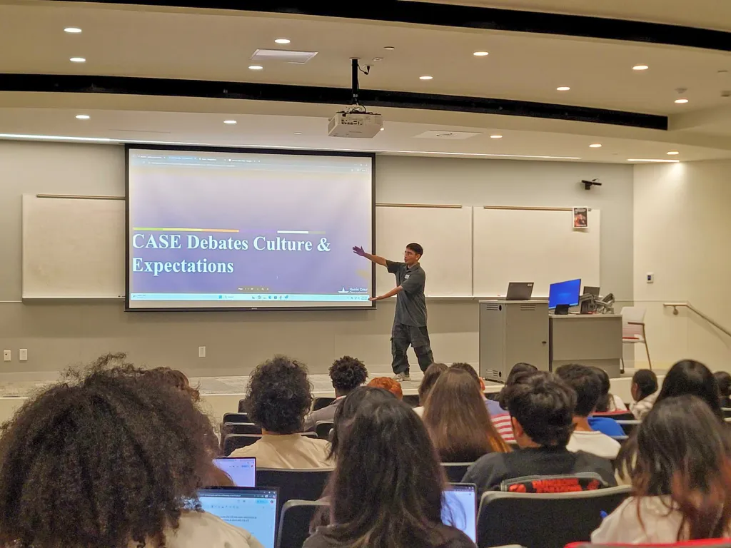 Instructor presenting to students in a tiered lecture hall, gesturing toward a projection screen