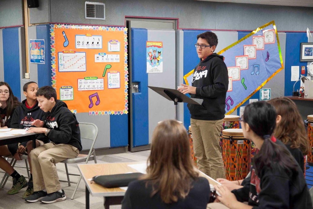 Student standing at a stand addressing peers seated at tables in a classroom, in a small-group instructional setting