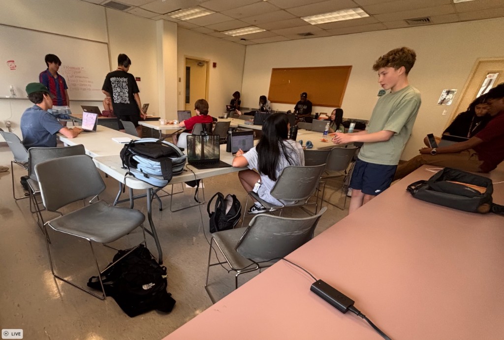 Students working at laptops along long tables in a bright classroom or computer lab, with a whiteboard in the background
