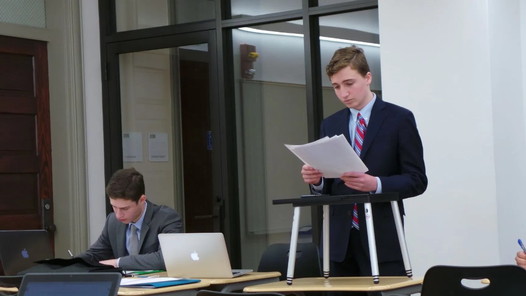 Students in formal attire: one standing at a lectern with notes, one seated at a desk with laptops