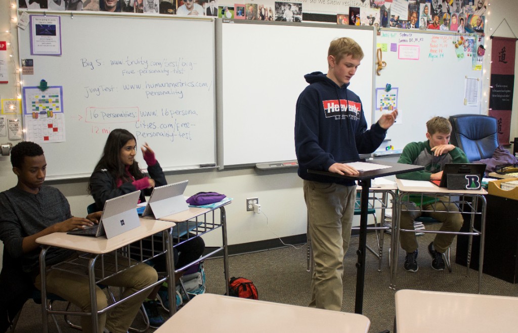 Students in a classroom with a speaker at a lectern and peers working on laptops